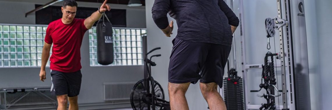 Back view of muscular man in sportswear doing exercises with trainer in modern fitness club